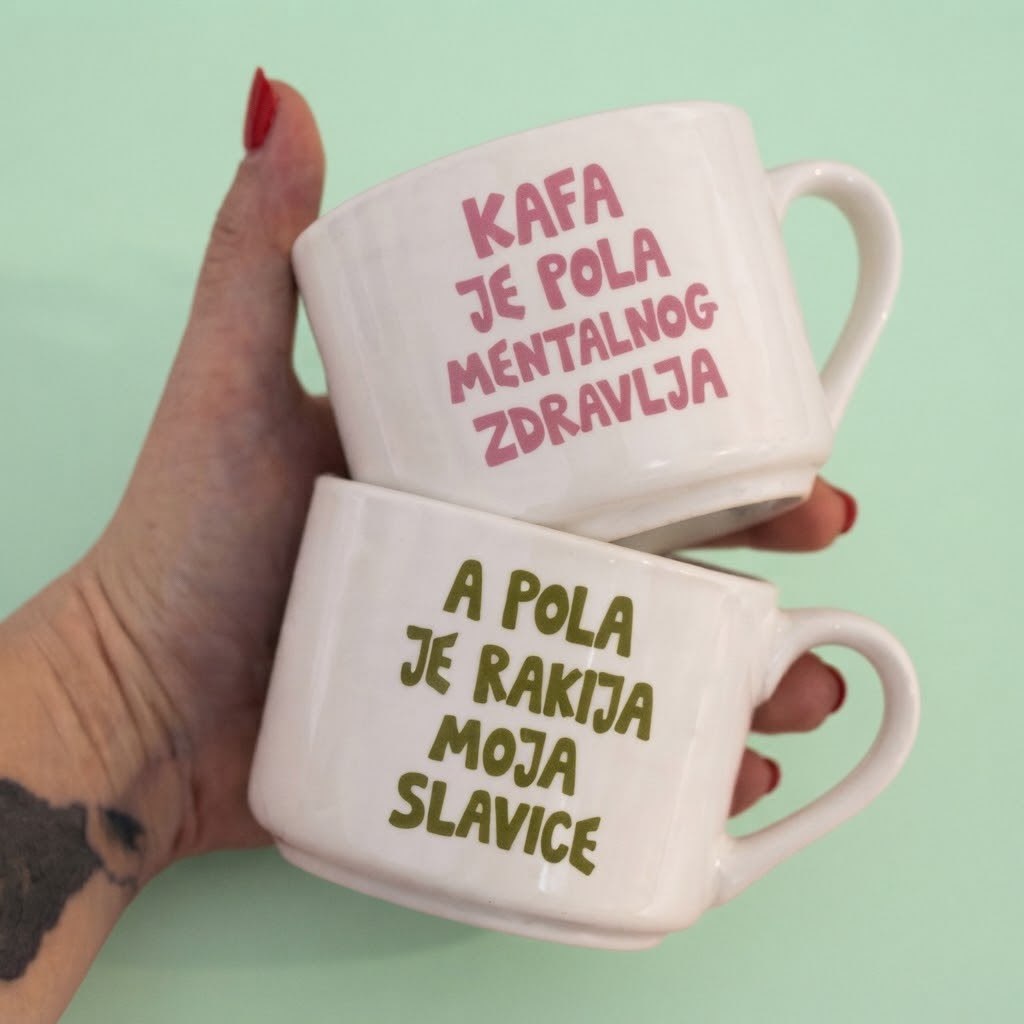 Dve bele šolje u ruci, natpisi Kafa je pola mentalnog zdravlja i A pola je rakija moja Slavice